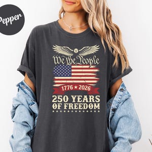 May include: Dark grey t-shirt with an American flag design and the text "We the People", "1776-2026", and "250 YEARS OF FREEDOM". The design includes an eagle. A casual wear item.