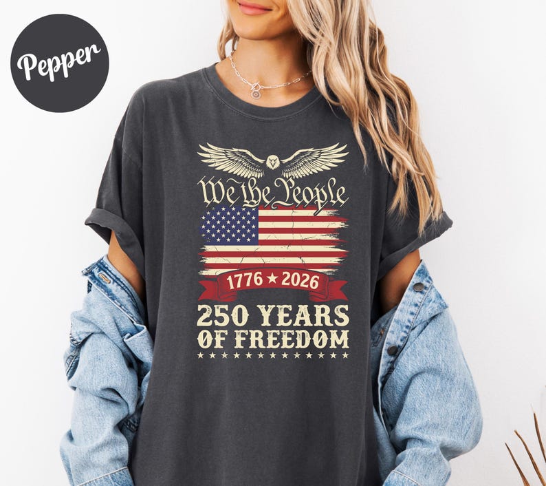 May include: Dark grey t-shirt with an American flag design and the text "We the People", "1776-2026", and "250 YEARS OF FREEDOM". The design includes an eagle. A casual wear item.