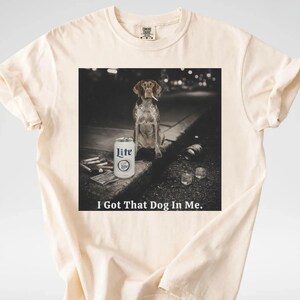 Pu&ograve; includere: T-shirt color crema con grafica di un cane con sigaretta, una lattina di birra Lite e la scritta "I Got That Dog In Me." L'immagine &egrave; in bianco e nero.