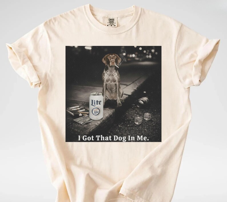 Pu&ograve; includere: T-shirt color crema con grafica di un cane con sigaretta, una lattina di birra Lite e la scritta "I Got That Dog In Me." L'immagine &egrave; in bianco e nero.