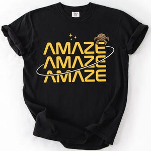 May include: Black t-shirt with the word "AMAZE" in yellow, repeated three times with a white curved line through the letters. A small turtle graphic and two yellow stars are above the word. The shirt is made of a soft material.