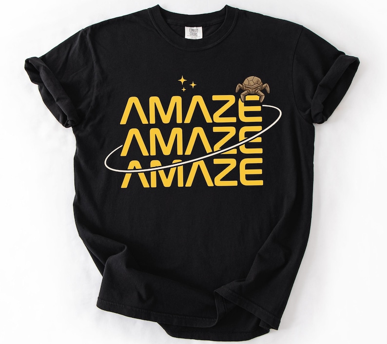 May include: Black t-shirt with the word "AMAZE" in yellow, repeated three times with a white curved line through the letters. A small turtle graphic and two yellow stars are above the word. The shirt is made of a soft material.