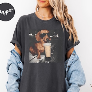 May include: Dark gray t-shirt featuring a dachshund wearing sunglasses and drinking an iced coffee. The dog is the focal point of a graphic print. The word "Pepper" is printed on a black circle in the upper left corner.