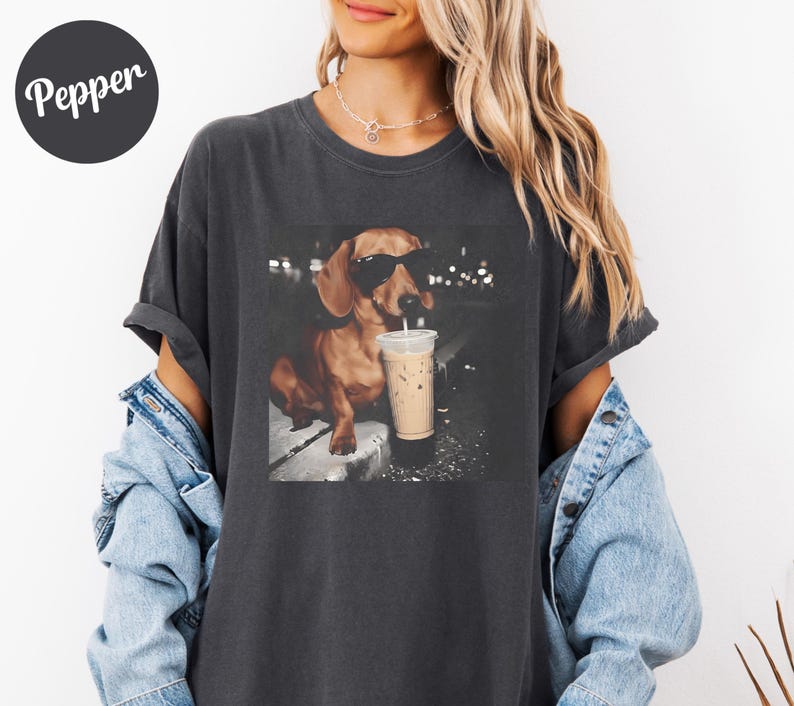 May include: Dark gray t-shirt featuring a dachshund wearing sunglasses and drinking an iced coffee. The dog is the focal point of a graphic print. The word "Pepper" is printed on a black circle in the upper left corner.