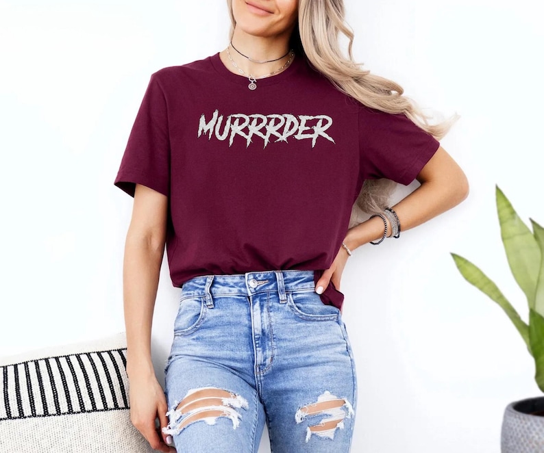May include: Burgundy t-shirt with the word "MURRRDER" in a distressed, white font. The shirt is a short-sleeved crew neck style. The model is wearing blue jeans with distressed patches.
