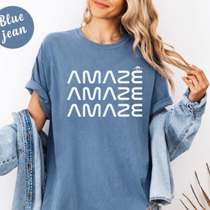 May include: A blue jean-colored t-shirt with the word "AMAZE" stacked three times in white. The shirt has short sleeves and a relaxed fit. A denim jacket is draped over the person's shoulders.