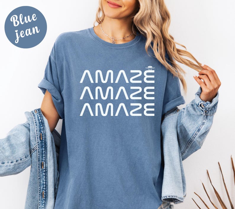 May include: A blue jean-colored t-shirt with the word "AMAZE" stacked three times in white. The shirt has short sleeves and a relaxed fit. A denim jacket is draped over the person's shoulders.