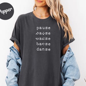 Puede incluir: Camiseta gris oscuro con la palabra "Pepper" en un círculo en la parte superior izquierda. La camiseta tiene la palabra "pause" y la palabra "danse" en blanco, con las letras entre las palabras cambiando.