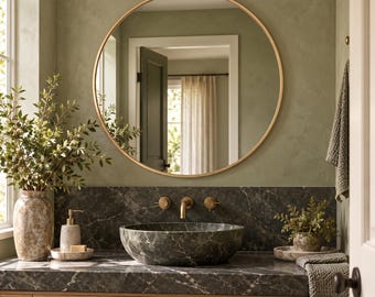 May include: A circular wall mirror with a thin gold frame hangs above a dark green marble vessel sink and countertop. The bathroom features a light wood vanity, a textured stone soap dispenser, and a green ceramic vase with leafy branches.