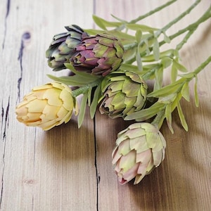 Puede incluir: Tallos de alcachofas artificiales en varios colores. Las alcachofas falsas son en tonos de verde, morado y amarillo, con hojas verdes. Los tallos están dispuestos sobre una superficie de madera, creando una exhibición natural y decorativa.