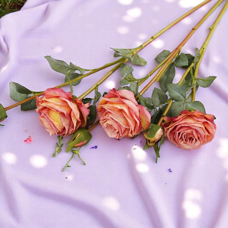 May include: Three artificial rose stems with peach and yellow flowers and green leaves on a light purple fabric background.