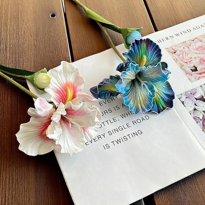 May include: Two artificial flower stems, one with white and pink flowers and the other with blue and iridescent flowers. The flowers are arranged on a white surface with text that reads "EVERY SINGLE ROAD IS TWISTING".