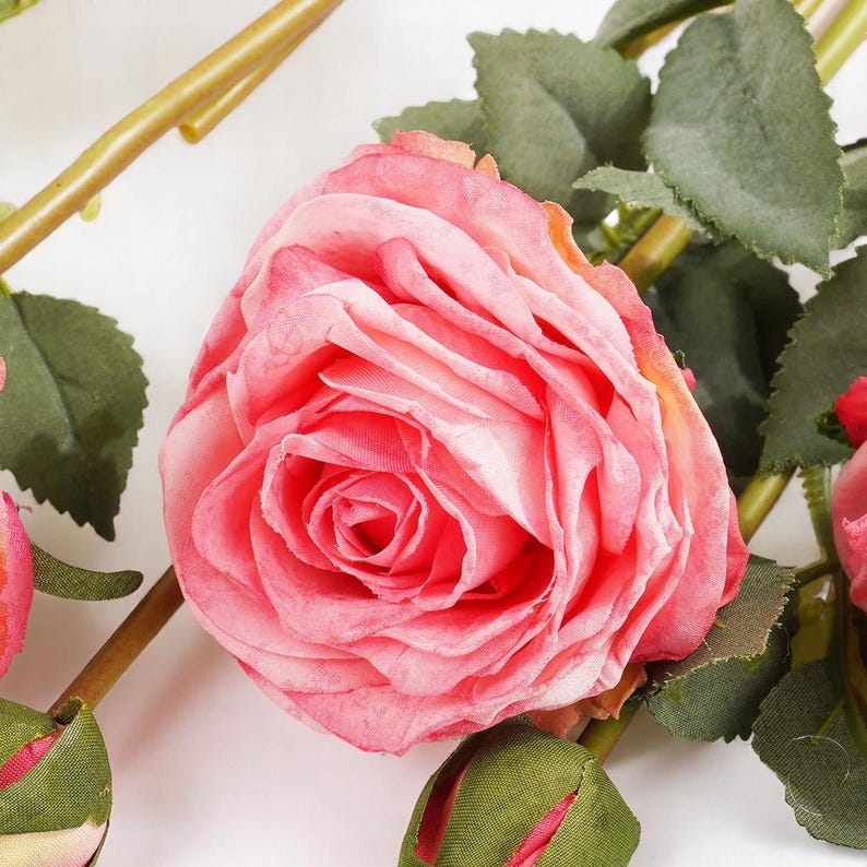 May include: A close-up of a pink artificial rose with green leaves. The rose is made of fabric and has a realistic appearance.
