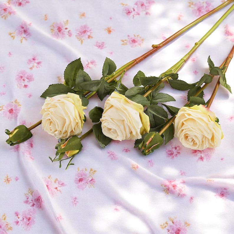 May include: Three cream-colored artificial roses with green leaves and brown stems on a white background with a pink floral pattern.