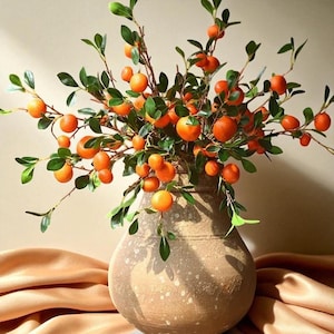 Op de afbeelding: Een decoratief bloemstuk met helder oranje fruit en groene bladeren. Gepresenteerd in een getextureerde, lichtbruine vaas. De vaas staat op een gedrapeerde, goudkleurige stof, wat een warme en uitnodigende uitstraling creëert.