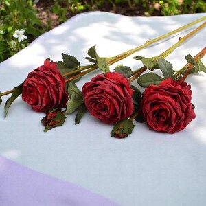 May include: Three artificial red roses with green leaves and stems on a white and purple surface.