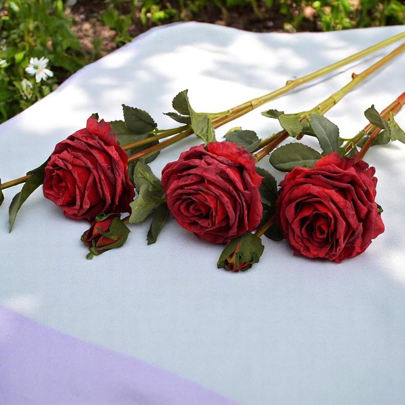 May include: Three artificial red roses with green leaves and stems on a white and purple surface.