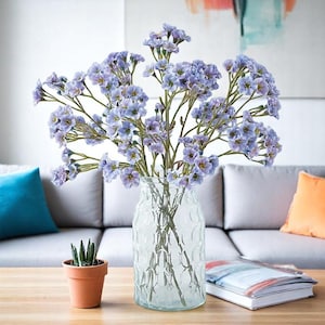 Puede incluir: Un jarrón de vidrio transparente con un patrón texturizado contiene un ramo de flores de color púrpura claro. Las flores tienen pequeños pétalos delicados y están dispuestas de forma natural. El jarrón está sobre una mesa de madera, con una pequeña planta en maceta y un libro cerca.