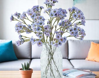 May include: A clear glass vase with a textured pattern holds a bouquet of light purple flowers. The flowers have small, delicate petals and are arranged in a full, natural style. The vase sits on a wooden table, with a small potted plant and a book nearby.