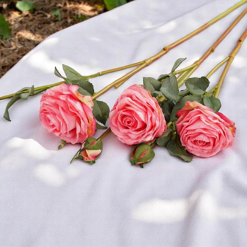 May include: Three pink artificial roses with green leaves and stems on a white fabric background.