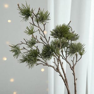 May include: Artificial evergreen pine branch with multiple sprigs of green needles and brown stems. The faux plant is a decorative accent for home decor, adding a touch of nature to any space.
