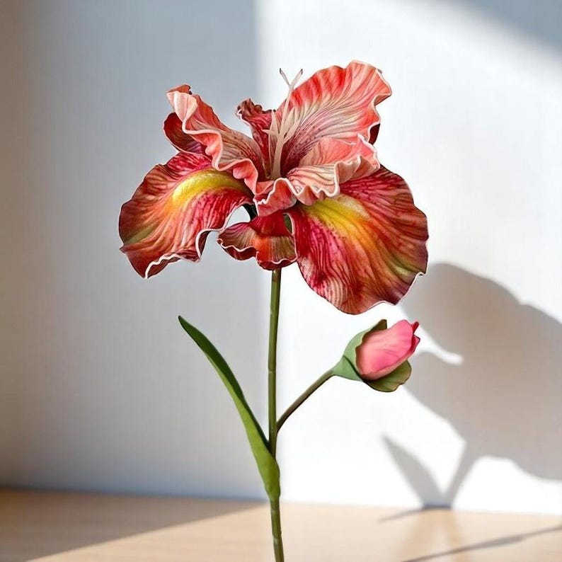 May include: A single artificial iris flower with a pink bud. The flower has red and yellow stripes and white edges. The stem is green.