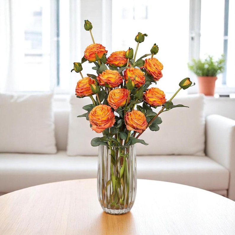 May include: A bouquet of orange and yellow artificial roses in a clear glass vase with vertical lines. The vase is sitting on a wooden table.