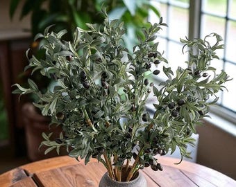 May include: An olive branch arrangement in a textured grey vase. The branches are filled with green leaves and dark purple olives, creating a natural aesthetic. The vase sits on a wooden table.