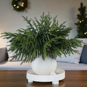 May include: A white, round vase with a textured surface, filled with green, leafy branches, sits on a white tray with short legs. A small, decorative ball rests on the tray. A Christmas wreath and small tree are in the background.