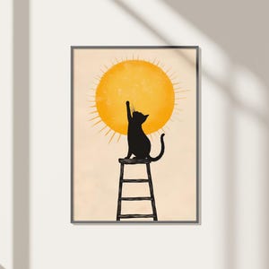 Könnte beinhalten: Gerahmter Kunstdruck mit einer schwarzen Katzen-Silhouette, die nach einer gelben Sonne greift. Die Katze sitzt auf einem kleinen, schwarzen Hocker. Der Hintergrund ist hellbeige, und das Kunstwerk ist in einem einfachen schwarzen Rahmen gerahmt.