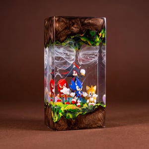 May include: A rectangular resin sculpture featuring Sonic the Hedgehog and friends. The scene includes Sonic, Tails, Knuckles, and Shadow figures set against a backdrop of a green and brown landscape with white cloud-like swirls. The sculpture is encased in a clear resin.