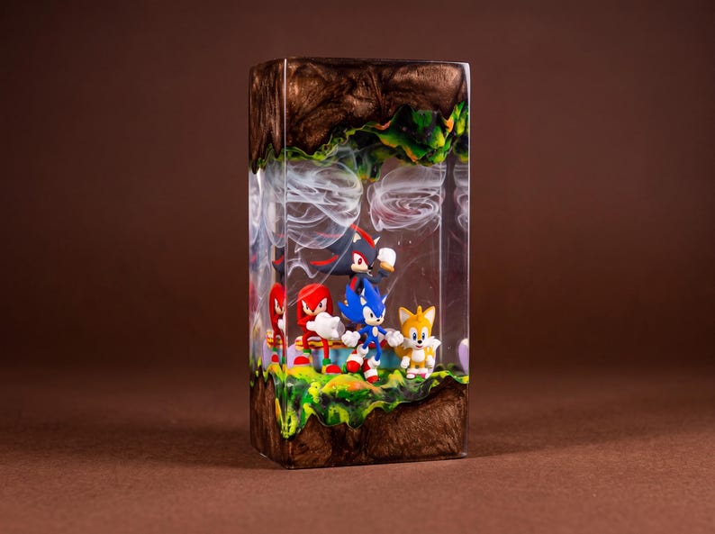 May include: A rectangular resin sculpture featuring Sonic the Hedgehog and friends. The scene includes Sonic, Tails, Knuckles, and Shadow figures set against a backdrop of a green and brown landscape with white cloud-like swirls. The sculpture is encased in a clear resin.