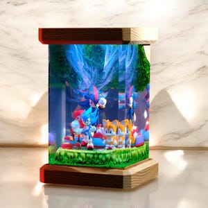 May include: A rectangular, illuminated display featuring Sonic the Hedgehog characters. The display has a wooden base and top with a clear acrylic box. Inside, Sonic, Tails, and Shadow figures stand on a green, grassy base, with a blue and white background.