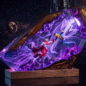 May include: A decorative resin and wood display piece featuring two superhero action figures in a dynamic combat pose. The scene is encased in clear resin with swirling purple smoke effects and jagged purple rock formations, set on a rectangular wooden base.