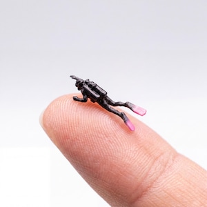 May include: A miniature black scuba diver figurine with pink flippers. The diver is lying on a fingertip, showcasing the small scale of the detailed diving equipment.