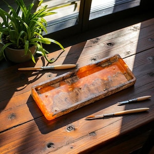 Puede incluir: Una bandeja rectangular de madera con un vibrante centro de resina naranja, acentuada con pequeños tachuelas color latón. Varios pinceles de artista con mangos de madera y virolas de metal están dispuestos alrededor de la bandeja, lo que sugiere un tema de artesanía o arte. La bandeja está sobre una superficie de madera.