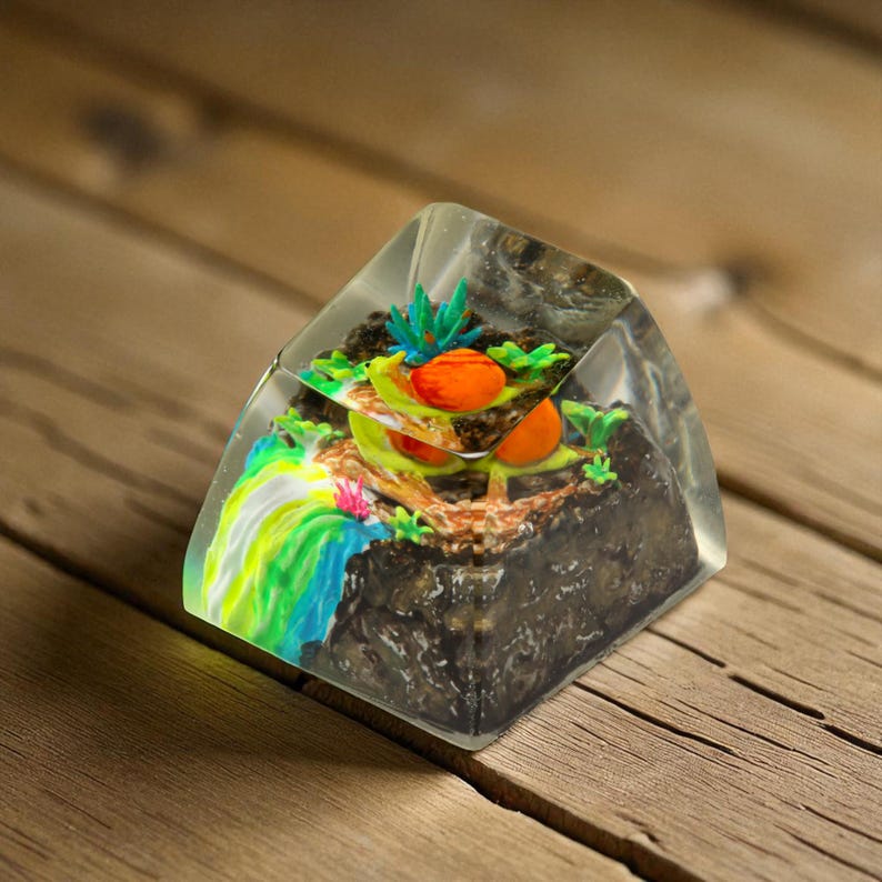 May include: A clear resin paperweight with a miniature landscape scene. The scene includes a waterfall with blue, green, and yellow hues, orange fruit, and green plants. The base is dark brown, resembling soil.