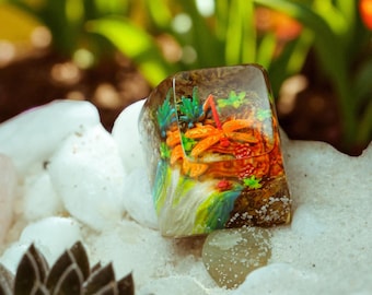 May include: A rectangular resin ring with a miniature underwater scene. The ring features orange, green, and brown elements, including what appears to be coral and a dragonfly. The ring is set against a backdrop of white stones and sand.