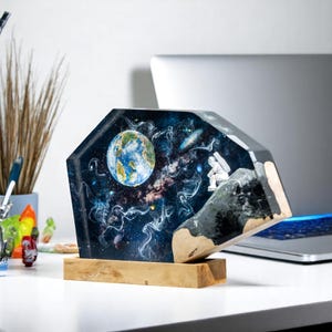 May include: A resin sculpture of an astronaut sitting on a moon-like surface with a view of Earth and a starry sky. The sculpture is on a wooden base.