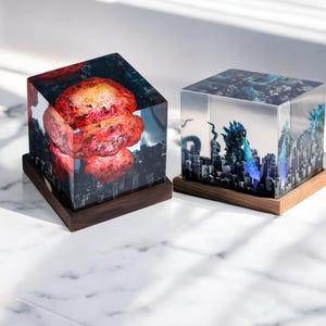 May include: Two resin art pieces. One cube features a red and orange mushroom cloud over a cityscape. The other cube depicts a blue monster attacking a city. Both are set on wooden bases.