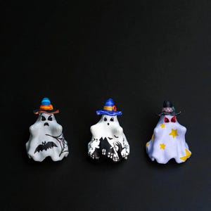 May include: Three Halloween ghost figurines on a black background. Each ghost wears a different colored witch hat. The ghosts are white with different designs, including a bat, a haunted house, and yellow stars.