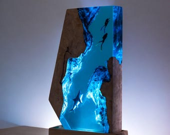Könnte beinhalten: Eine einzigartige Holzlampe mit blauem Harzkern, die Silhouetten von Tauchern und einen Hai zeigt. Die Lampe hat eine rechteckige Form mit einem Holzsockel und einem Licht, das das Harz beleuchtet und eine faszinierende Unterwasserszene erzeugt.
