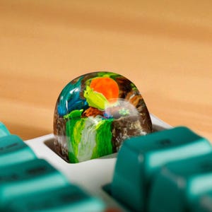 May include: A custom keycap with a colorful, artistic design. The keycap features a landscape with a yellow bird, orange sun, and green waterfall. The keycap is set in a white tray with teal keys.