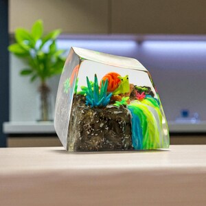 May include: A clear resin paperweight featuring a miniature underwater scene. It includes a colorful snail, coral, and aquatic plants. The base is a dark, textured rock formation, with a rainbow-colored waterfall effect on the side.