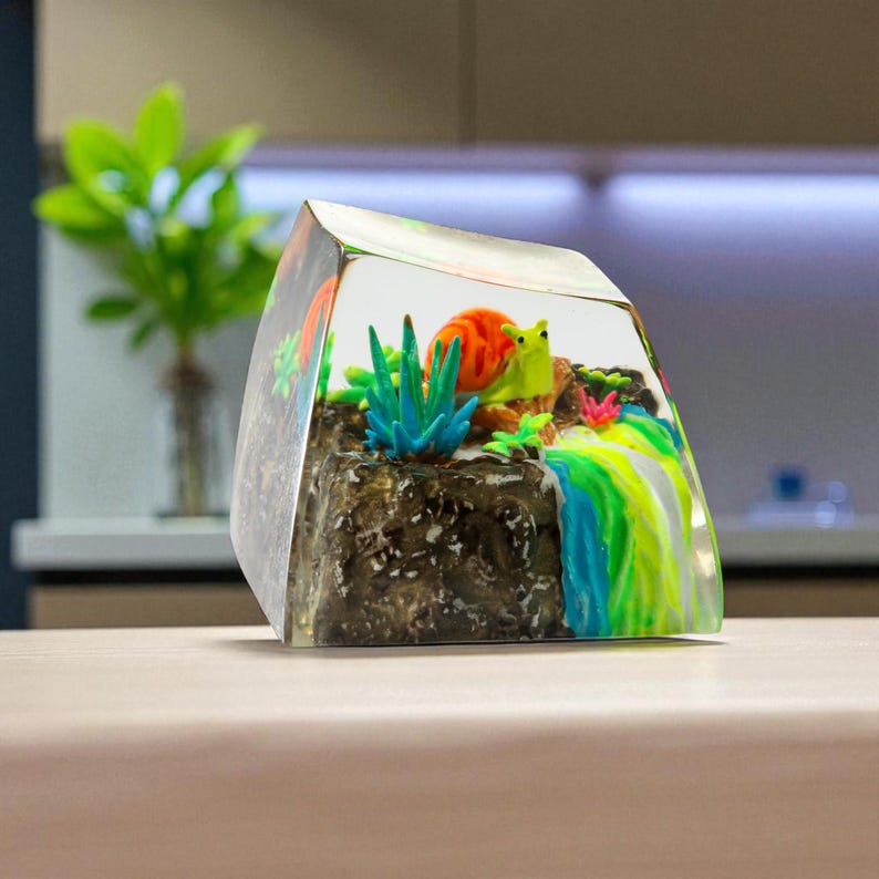 May include: A clear resin paperweight featuring a miniature underwater scene. It includes a colorful snail, coral, and aquatic plants. The base is a dark, textured rock formation, with a rainbow-colored waterfall effect on the side.