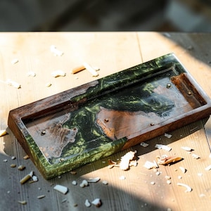 May include: A rectangular ashtray with a wooden frame and a green and brown resin interior. The ashtray has a smooth, polished finish and small brass accents. It sits on a wooden surface, with cigarette remnants scattered around.
