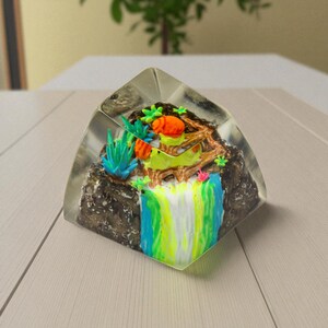 May include: A clear, triangular resin paperweight with a miniature landscape. It features a waterfall with blue, green, and yellow colours, a brown rock formation, and colourful plants. The paperweight is set on a light-coloured wooden surface.