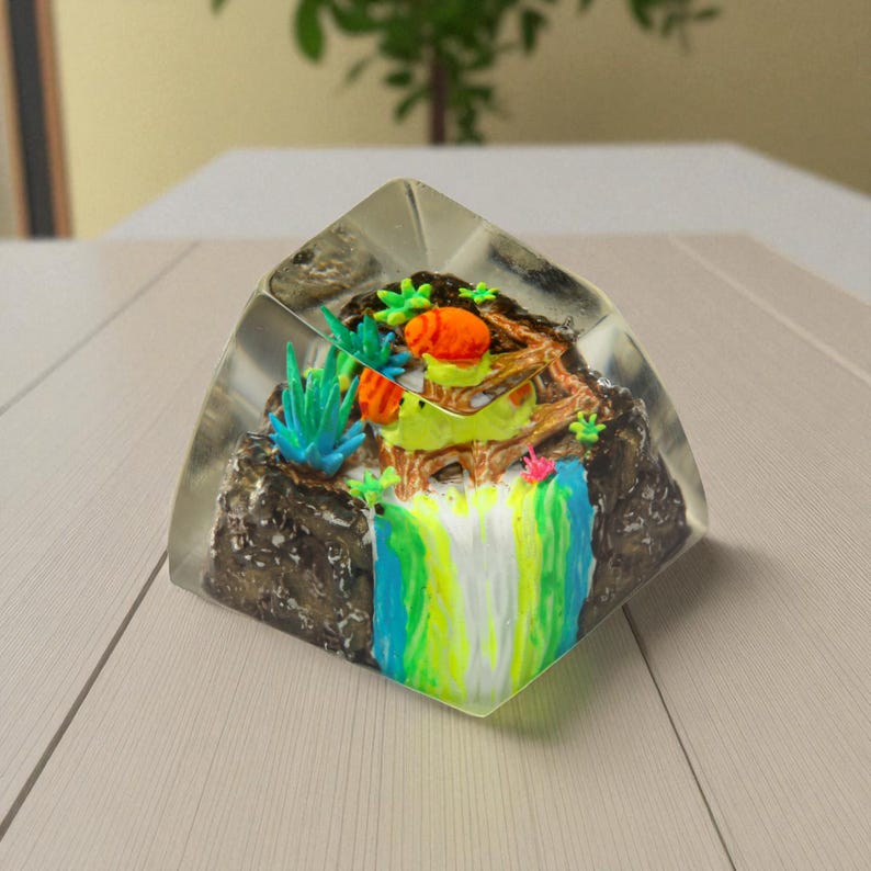 May include: A clear, triangular resin paperweight featuring a miniature landscape. The scene includes a waterfall with blue, green, and yellow hues, a brown rock formation, and colorful plants. The paperweight is set on a light-colored wooden surface.