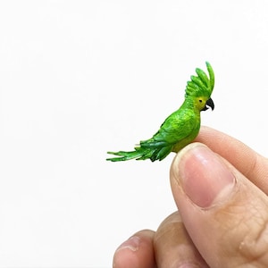 Puede incluir: Una figura de loro verde en miniatura con una cresta y plumas de la cola detalladas. El pequeño pájaro tiene un pico negro y se sostiene entre dos dedos sobre un fondo blanco. El loro es de un tono verde vibrante.