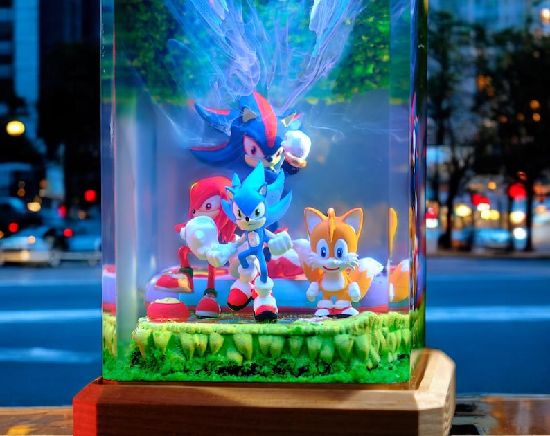 May include: A clear, rectangular display case featuring Sonic the Hedgehog and friends. The figures include Sonic, Knuckles, Tails, and Shadow, set on a green base. The case is mounted on a wooden base, with a city street visible in the background.